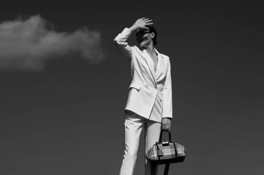 Beyaz takım elbiseli kız elini yüzünü kapatıyor, gökyüzüne karşı duruyor çanta, siyah beyaz fotoğraf, derin gölgeler