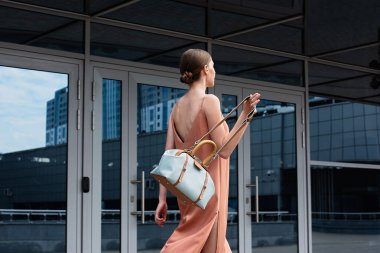 Elinde çanta olan bir kız binanın arka planında yürüyor, camlarda yansıma var.