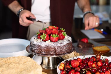 Çikolatalı kek mutfaktaki masada duruyor, tatlılık çilekle süsleniyor, arka planda bıçaklarla, seçici bir odak ile.