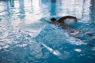 Havuzda yüzen atletik bir adam, mavi su, sudaki pencerelerin yansıması, insan hareketinden yayılan su spreyi, seçici odaklanma.