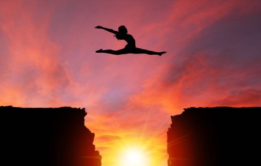 Silhouette of girl dancer in a split leap over dangerous cliffs with dramatic sunset or sunrise background and copy space. Concept of faith, conquering adversity, taking risk; challenge, courage, determination or achievement. clipart