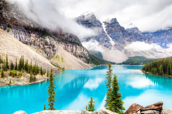Fog and clouds descend onto the Valley of Ten Peaks where glacier-fed Moraine Lake gives off its distinct turquoise blue color due to refraction of light off the rock flour at the bottom of the lake.