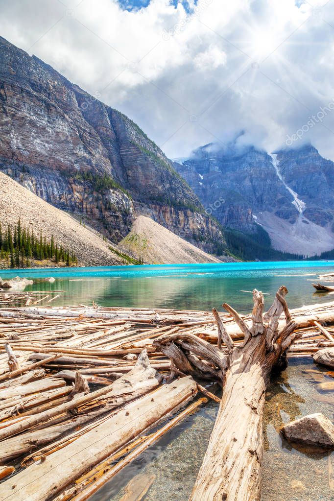 Driftwood on the shores of Moraine Lake with sun peeking through the clouds overshadowing the mountains of Valley of Ten Peaks.