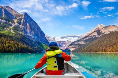 Teen boy canoeing on Lake Louise in Banff National Park of the Canadian Rockies with its glacier-fed turquoise lakes and Mount Victoria Glacier in the background. clipart