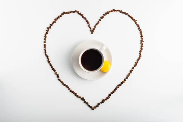 Heart formed from coffee beans surrounds a cup of hot coffee and yellow macaron on a plate, top view