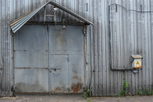 Kapalı gri kapısı olan eski bir metal endüstriyel binanın ön cephesinin bir parçası, üstünde bir kamera ve duvarında bir elektrik kutusu var.