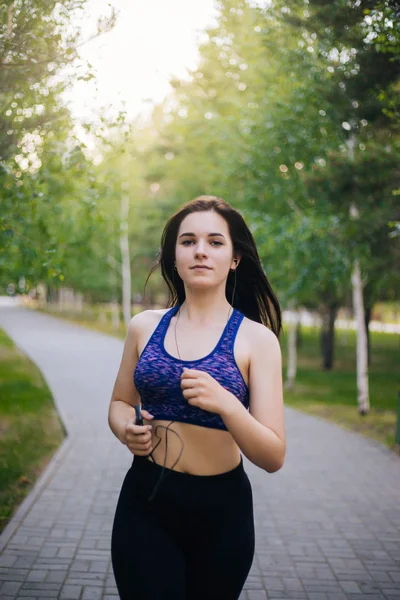 Young beautiful girl on a run. A woman is engaged in sports in the park. Healthy lifestyle. Running along the path with headphones, listening to music.