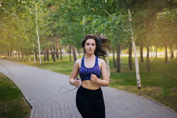 Young beautiful girl on a run. A woman is engaged in sports in the park. Healthy lifestyle. Running along the path with headphones, listening to music.