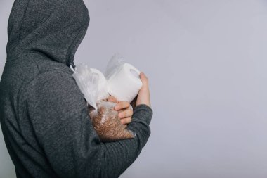 A guy in a gray sweatshirt on a light background in a medical mask holds essential products. A man in a panic buys toilet paper and buckwheat groats, rice.