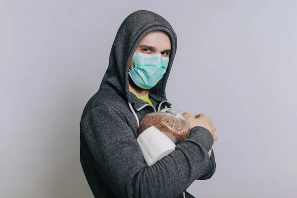 Un gars en sweat-shirt gris sur un fond clair dans un masque médical détient des produits essentiels. Un homme paniqué achète du papier toilette et des gruaux de sarrasin, du riz . — Photo