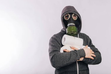 A guy in a gas mask and a gray sweatshirt in a hood holds rolls of toilet paper in his hands on a white background. The stink in the toilet. Panic during quarantine.