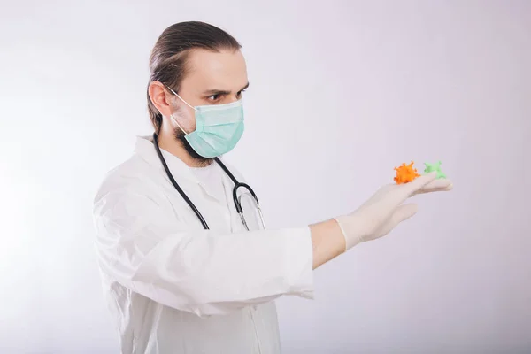 A doctor in a white coat and gloves on a white background holding a mock coronavirus in his hands. Shows a multi-colored virus. A male paramedic in a medical mask contracted a dangerous disease — Φωτογραφία Αρχείου