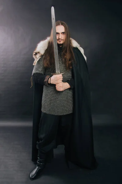 Warrior in armor with a sword. A guy with long hair and a beard, a fox collar. Dressed in chain mail, a black cloak and black pants holds a sword in his hands. Studio photo against a dark background. — ストック写真