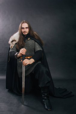 Warrior in armor with a sword. A guy with long hair and a beard, a fox collar. Dressed in chain mail, a black cloak and black pants holds a sword in his hands while sitting on one knee.