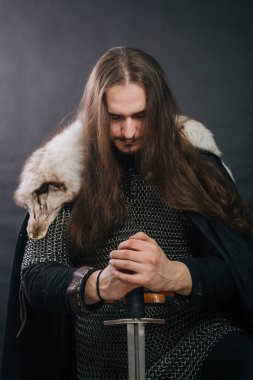 Warrior in armor with a sword. A guy with long hair and a beard, a fox collar. Dressed in chain mail, a black cloak and black pants holds a sword in his hands while sitting on one knee.