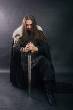 Warrior in armor with a sword. A guy with long hair and a beard, a fox collar. Dressed in chain mail, a black cloak and black pants holds a sword in his hands while sitting on one knee.