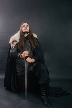 Warrior in armor with a sword. A guy with long hair and a beard, a fox collar. Dressed in chain mail, a black cloak and black pants holds a sword in his hands while sitting on one knee.