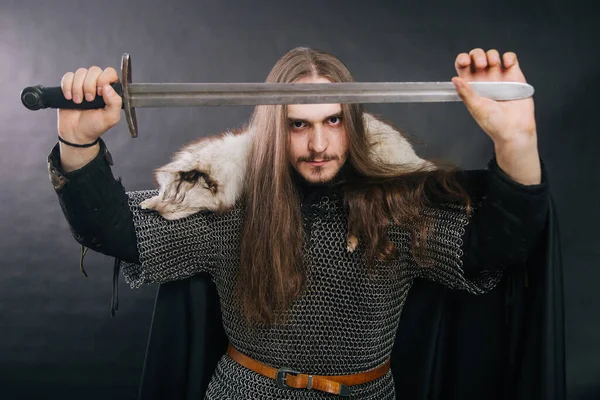 Guerrier en armure avec une épée. Un type aux cheveux longs et à la barbe, un collier de renard. Vêtu d'une chaîne de courrier, un manteau noir et un pantalon noir tient une épée dans ses mains . — Photo