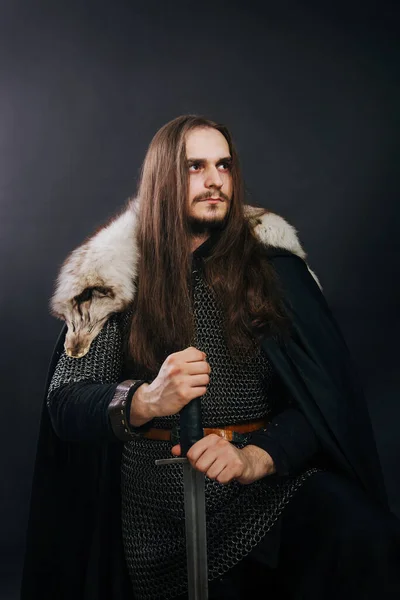 Knight in chain mail and with a fur collar on a black cloak on a gray background. Portrait of a Viking man with long hair and a beard in armor holds a sword.