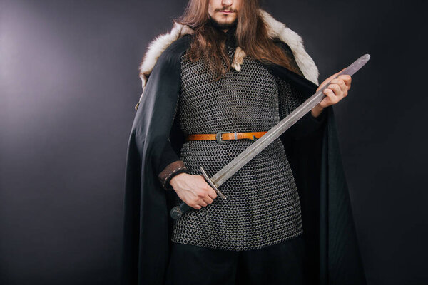 Knight in chain mail and with a fur collar on a black cloak on a gray background. Portrait of a Viking man with long hair and a beard in armor holds a sword.