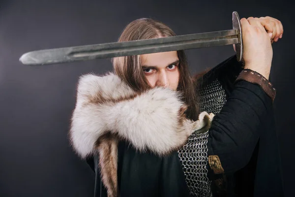 Knight in chain mail and with a fur collar on a black cloak on a gray background. Portrait of a Viking man with long hair and a beard in armor holds a sword.