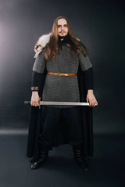 Warrior in armor with a sword. A guy with long hair and a beard, a fox collar. Dressed in chain mail, a black cloak and black pants holds a sword in his hands.