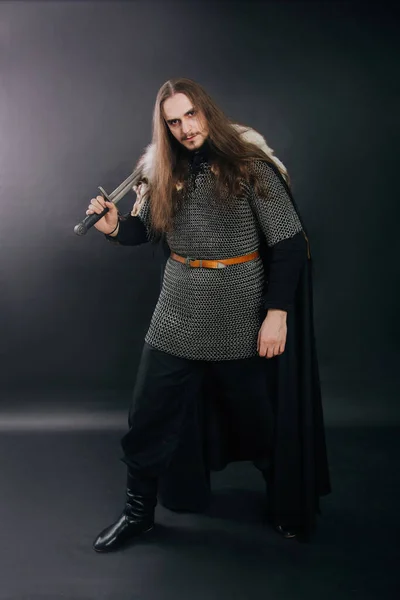 Warrior in armor with a sword. A guy with long hair and a beard, a fox collar. Dressed in chain mail, a black cloak and black pants holds a sword in his hands.