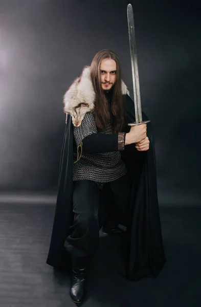 Warrior in armor with a sword. A guy with long hair and a beard, a fox collar. Dressed in chain mail, a black cloak and black pants holds a sword in his hands.