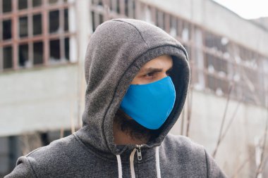 A guy in a gray sweater with a blue medical mask. Holds a hood with his own hands. Against the background of destroyed and abandoned buildings. Quarantined man.