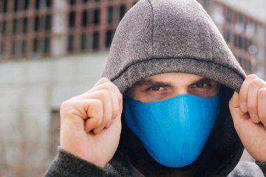 A guy in a gray sweater with a blue medical mask. Holds a hood with his own hands. Against the background of destroyed and abandoned buildings. Quarantined man.