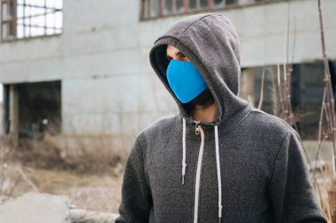 A guy in a gray sweater with a blue medical mask. Holds a hood with his own hands. Against the background of destroyed and abandoned buildings. Quarantined man.