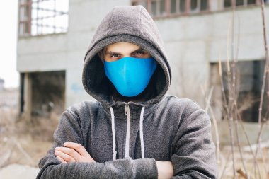 A guy in a gray sweater with a blue medical mask. Holds a hood with his own hands. Against the background of destroyed and abandoned buildings. Quarantined man.