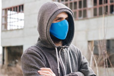 A guy in a gray sweater with a blue medical mask. Holds a hood with his own hands. Against the background of destroyed and abandoned buildings. Quarantined man.