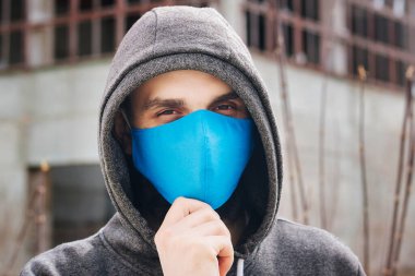 A guy in a gray sweater with a blue medical mask. Holds a hood with his own hands. Against the background of destroyed and abandoned buildings. Quarantined man.