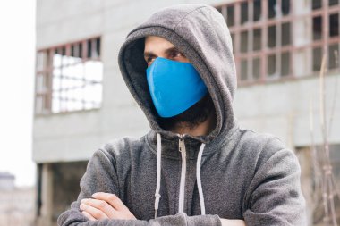 A guy in a gray sweater with a blue medical mask. Holds a hood with his own hands. Against the background of destroyed and abandoned buildings. Quarantined man.