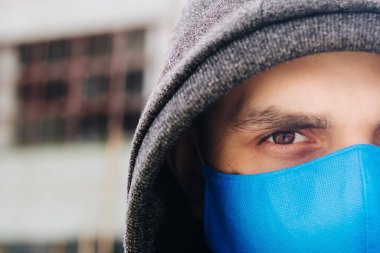 A guy in a gray sweater with a blue medical mask. Holds a hood with his own hands. Against the background of destroyed and abandoned buildings. Quarantined man.