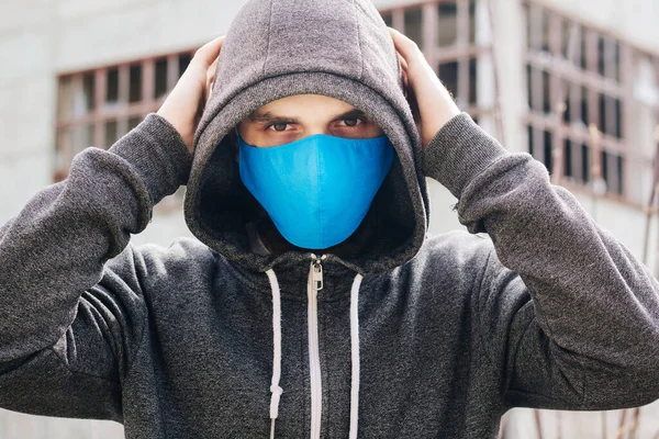 A guy in a gray sweater with a blue medical mask. Holds a hood with his own hands. Against the background of destroyed and abandoned buildings. Quarantined man.