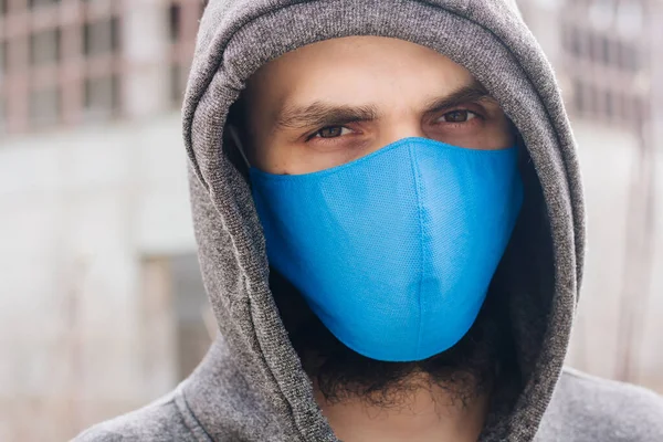 A guy in a gray sweater with a blue medical mask. Holds a hood with his own hands. Against the background of destroyed and abandoned buildings. Quarantined man.