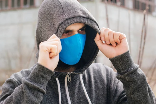 A guy in a gray sweater with a blue medical mask. Holds a hood with his own hands. Against the background of destroyed and abandoned buildings. Quarantined man.