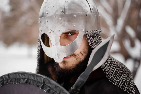 Viking in winter with a spear and a round shield of red-black color. A guy in a helmet and chain mail in the snow.