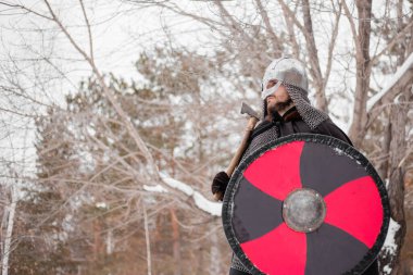 Viking in winter with an ax and a round shield in red-black. A guy in a helmet and chain mail in the snow.