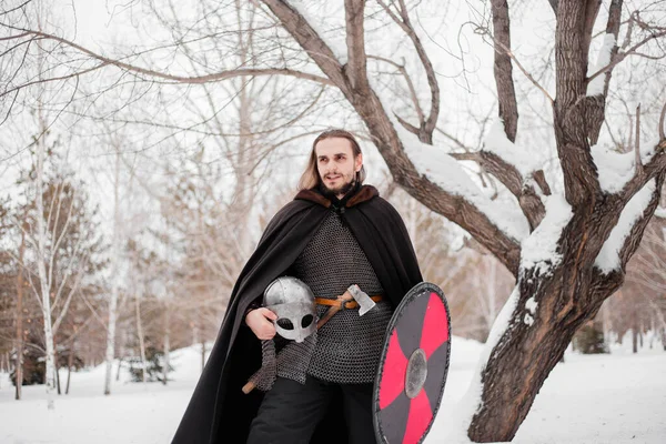 A handsome young guy with long hair and in armor holds a helmet in his hands. Warrior in a black cloak in the winter in the forest to prepare for battle.