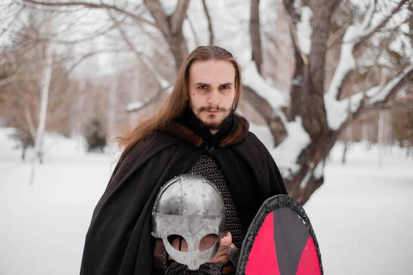 A handsome young guy with long hair and in armor holds a helmet in his hands. Warrior in a black cloak in the winter in the forest to prepare for battle.