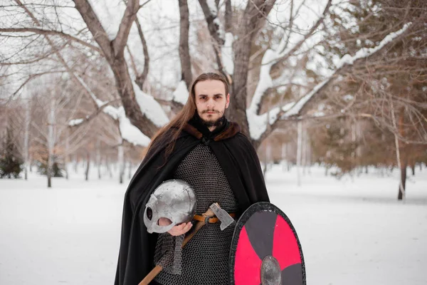 A handsome young guy with long hair and in armor holds a helmet in his hands. Warrior in a black cloak in the winter in the forest to prepare for battle.