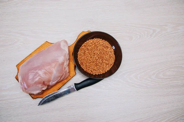 Ingredient for cooking. Raw turkey lies on a wooden board on a white kitchen table. Filet of the bird is ready for cutting with a knife. Dark cup with buckwheat.