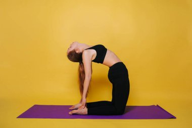Athletic, beautiful young girl practicing yoga doing ushtrasana pose. Wear black sportswear on a plain yellow background.