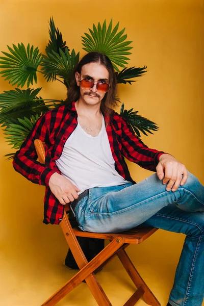 一个长头发 留着胡子 穿着80年代红色格子花迪斯科衬衫的有魅力的男人坐在椅子上 戴着时髦太阳镜的家伙 背景是一棵绿色棕榈树 — 图库照片