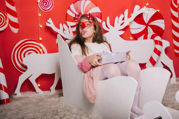 A cheerful girl in glasses with a red nose and horns is holding a gift in her hands. A wall of lollipops. The child is sitting in a Christmas sleigh.