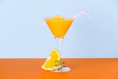 Taze portakal suyu bardakta bir dilim ve turuncu yüzeyde bir nane yaprağı ve açık mavi bir zemin.