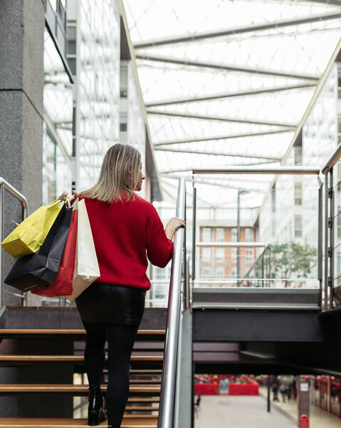 Adult woman wearing a red sweater walking up the stairs of a shopping mall carrying colorful shopping bags.